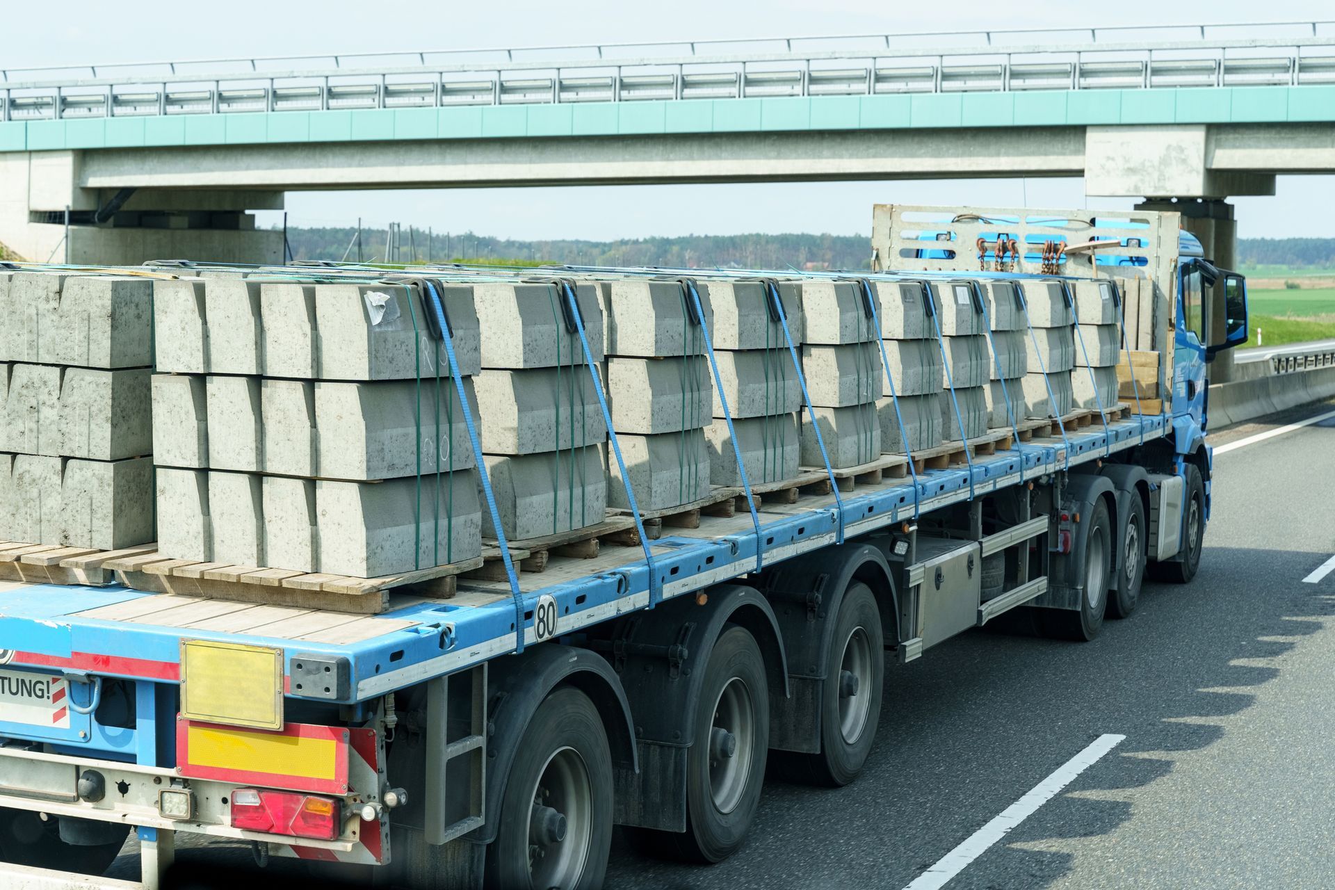 Flatbed truck carrying stacked concrete blocks on a highway, passing under a bridge.