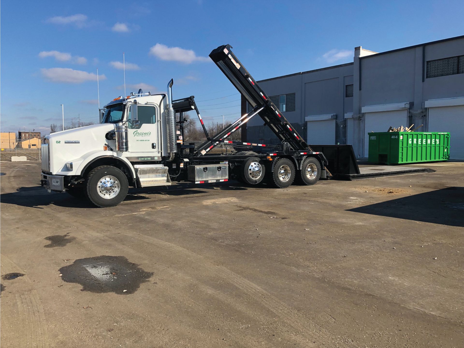 White roll-off truck lifting a dumpster in front of a gray building on a sunny day.