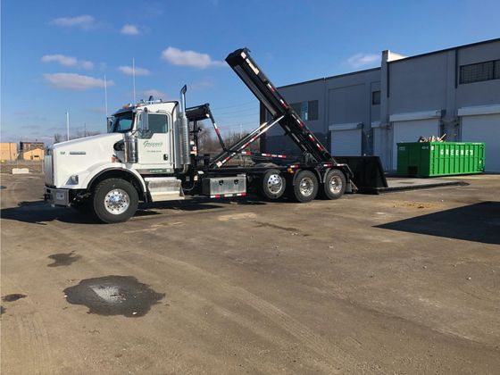 White roll-off truck lifting a dumpster in front of a gray building on a sunny day.