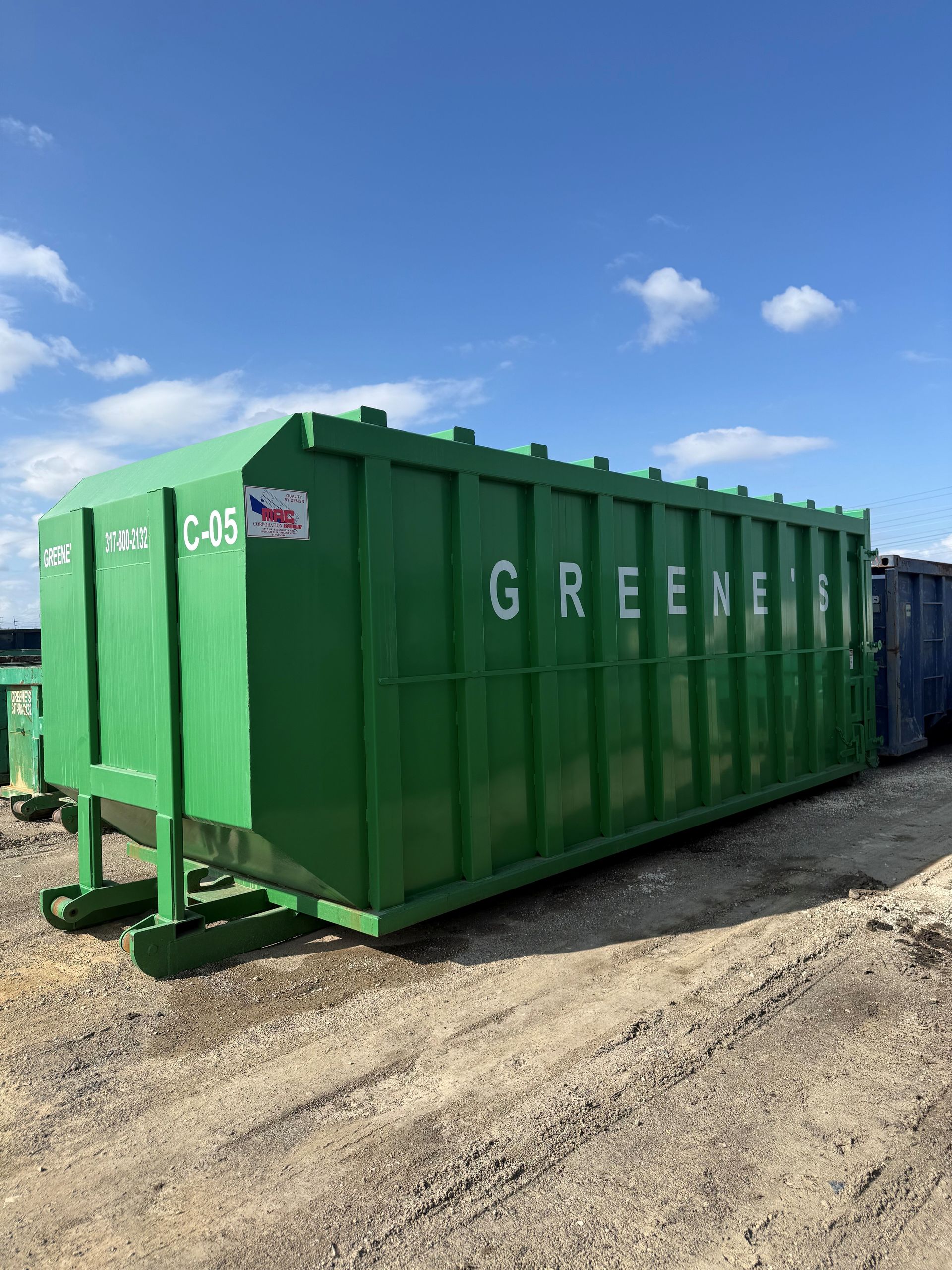 Three dumpsters, blue and green, next to a white car parked under a 'P' sign. Buildings with green roofs in background.