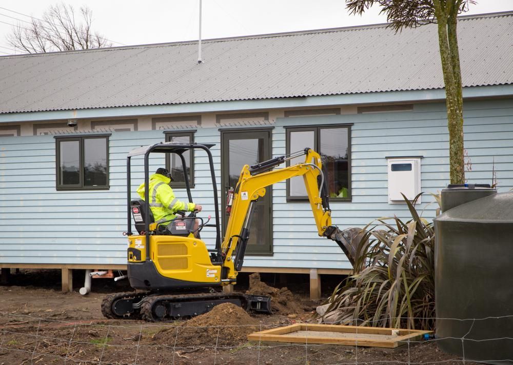 A Man is Driving a Small Yellow Excavator in Front of a House — Husky Demolition In Forster, NSW