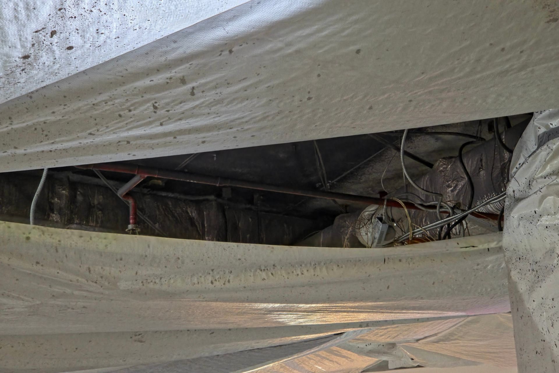 View from inside a crawl space, showing insulation, pipes, wiring, and dark space beyond.