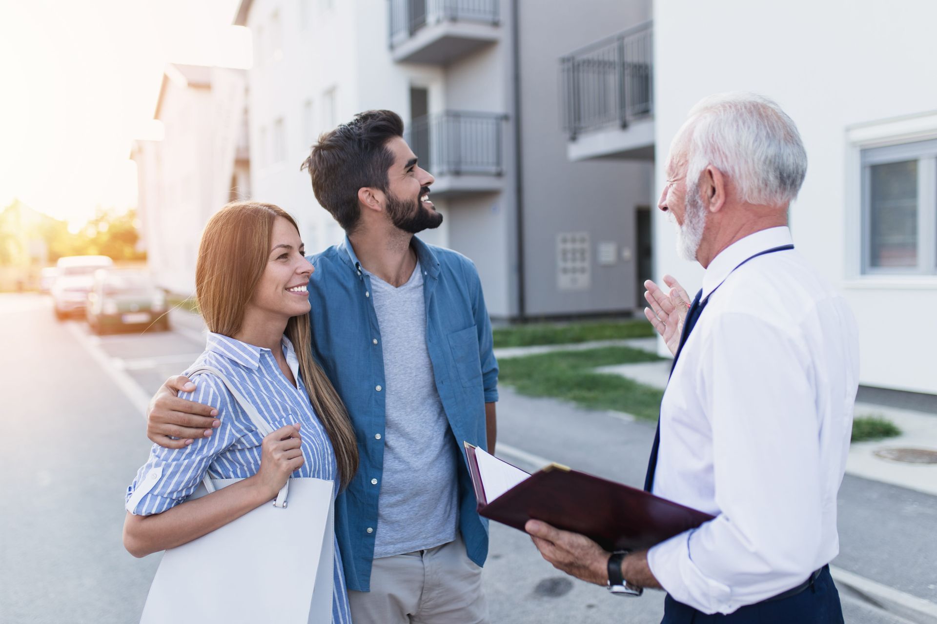 Couple with a realtor looking at apartments. They stand on a street, smiling, while he holds a folder.
