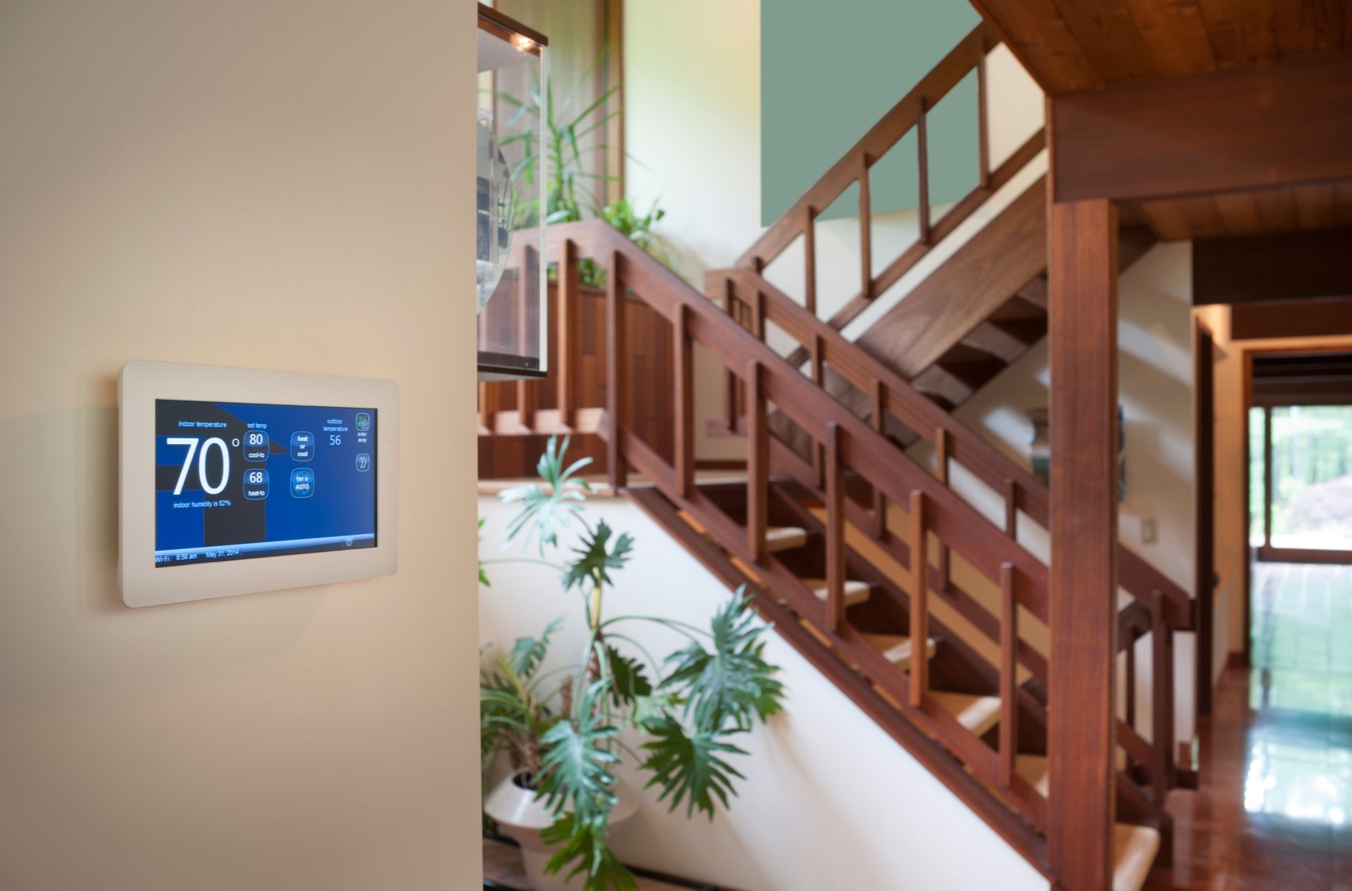 Smart thermostat displaying 70 degrees on a beige wall near a wooden staircase with potted plants.