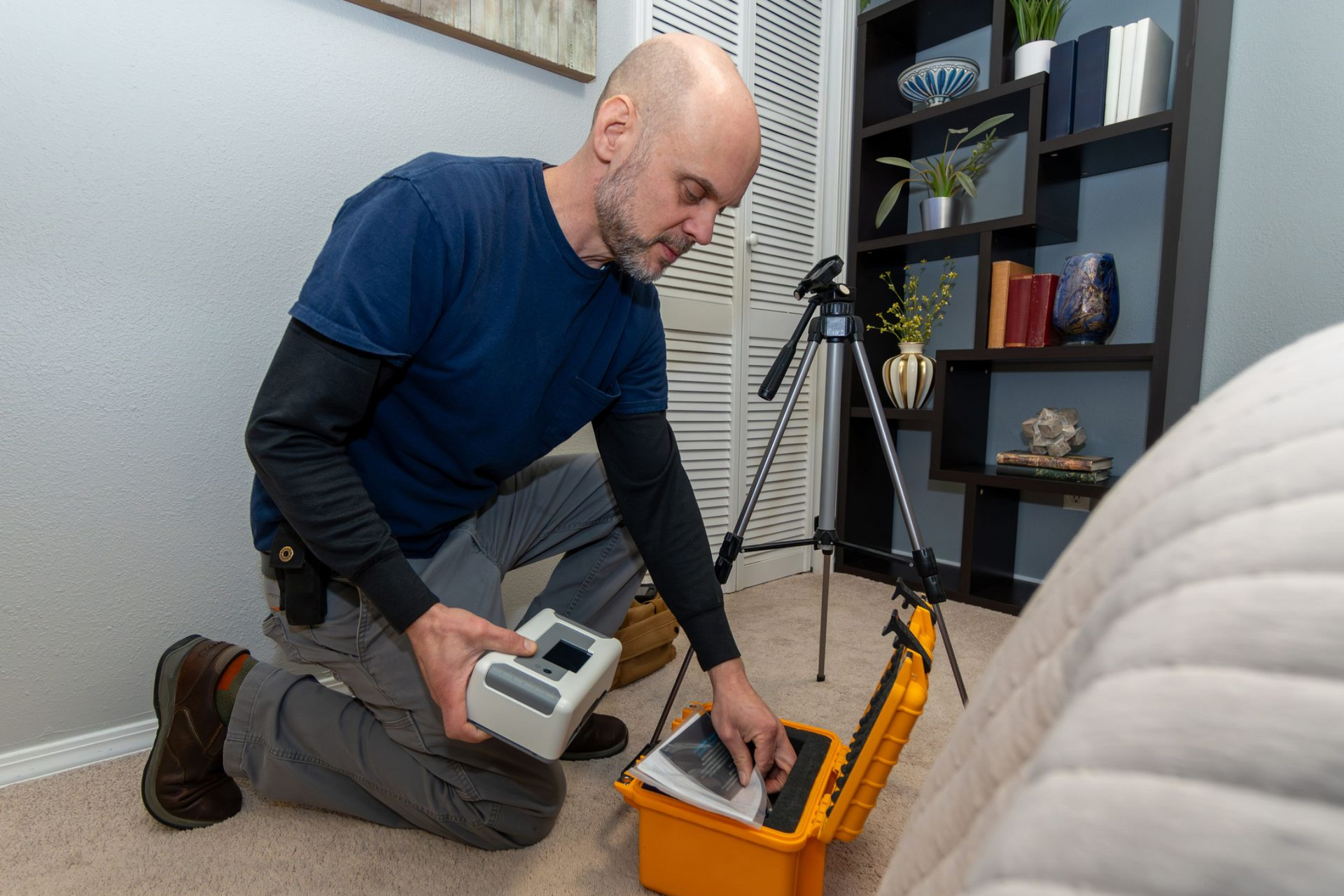 Man inspecting equipment in a case near a bed, a tripod, and a bookcase in a room.