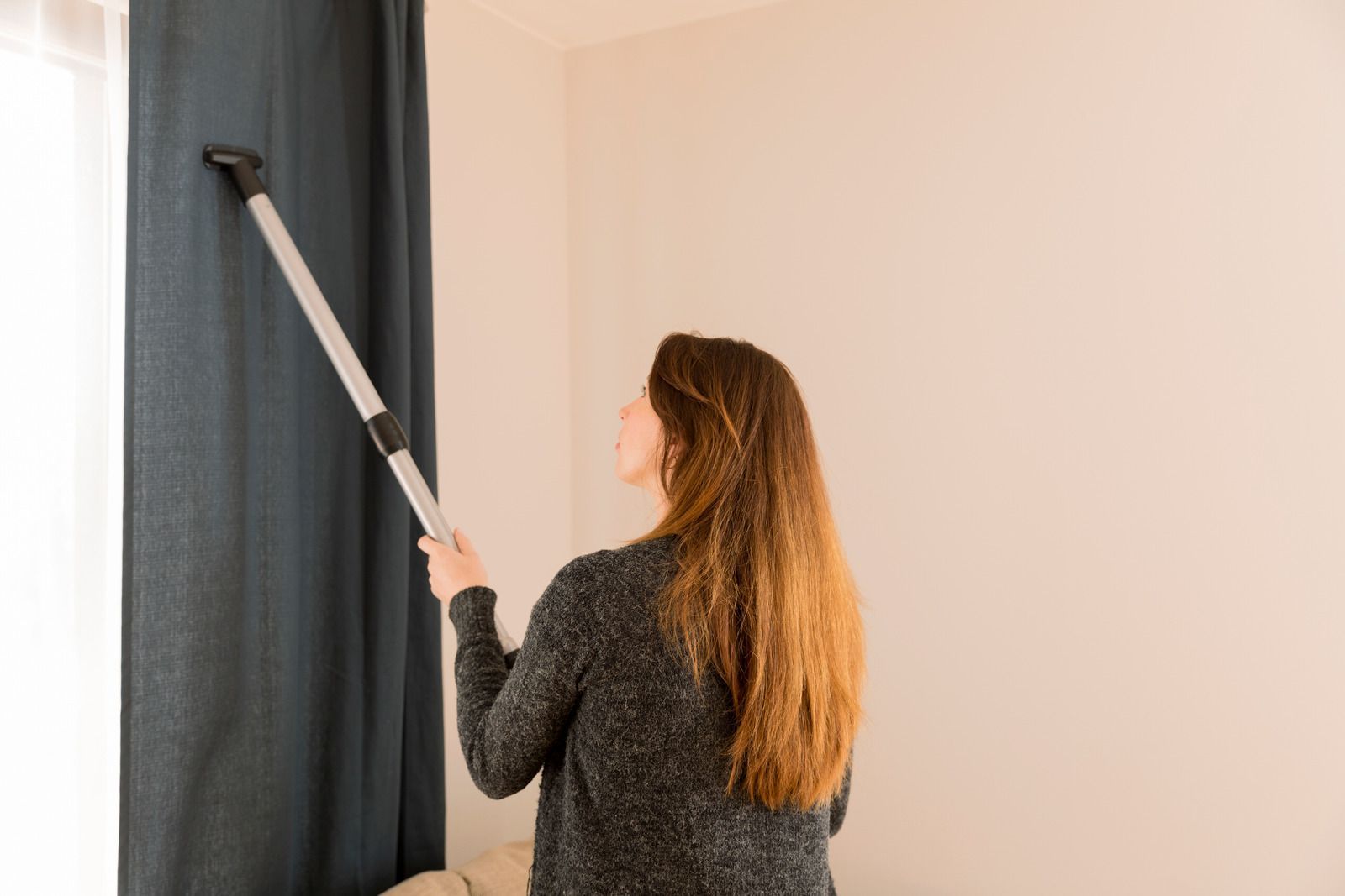 A woman is cleaning curtains with a vacuum cleaner.