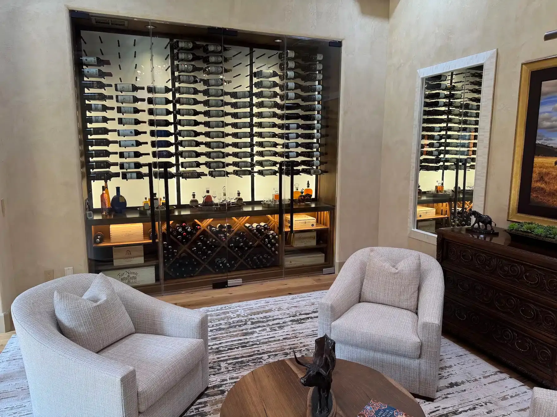 Modern wine cellar with wall-mounted racks, two white chairs, a rug, and a wood cabinet with a mirror.