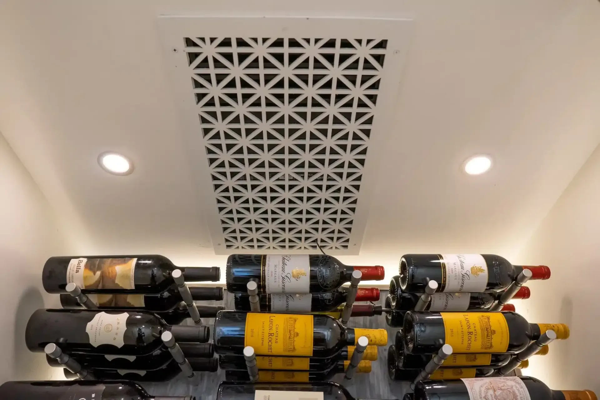 Wine bottles stacked in a rack beneath a decorative ceiling vent and recessed lights