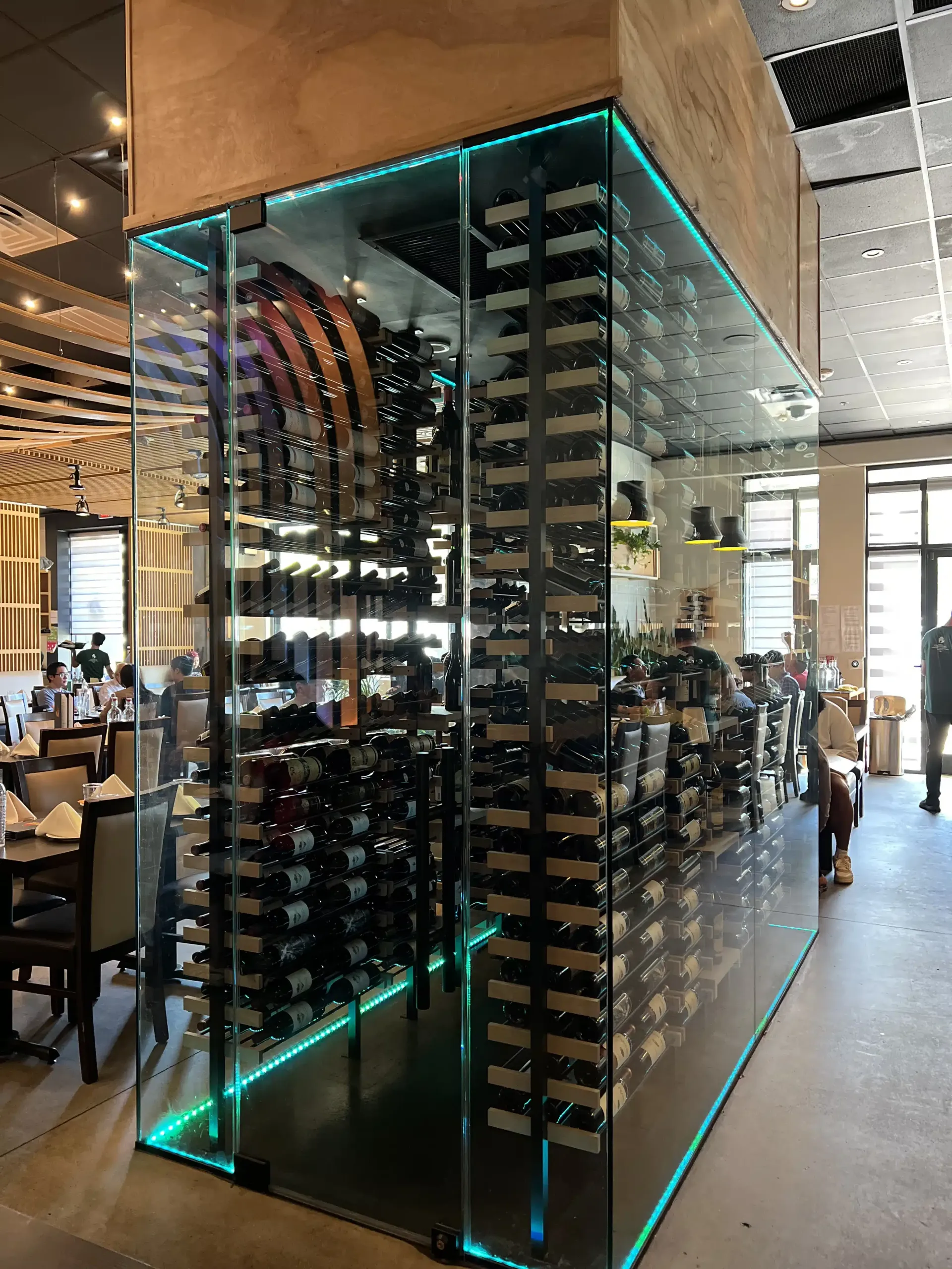 Glass wine display case with black shelving and turquoise LED trim in a modern restaurant.
