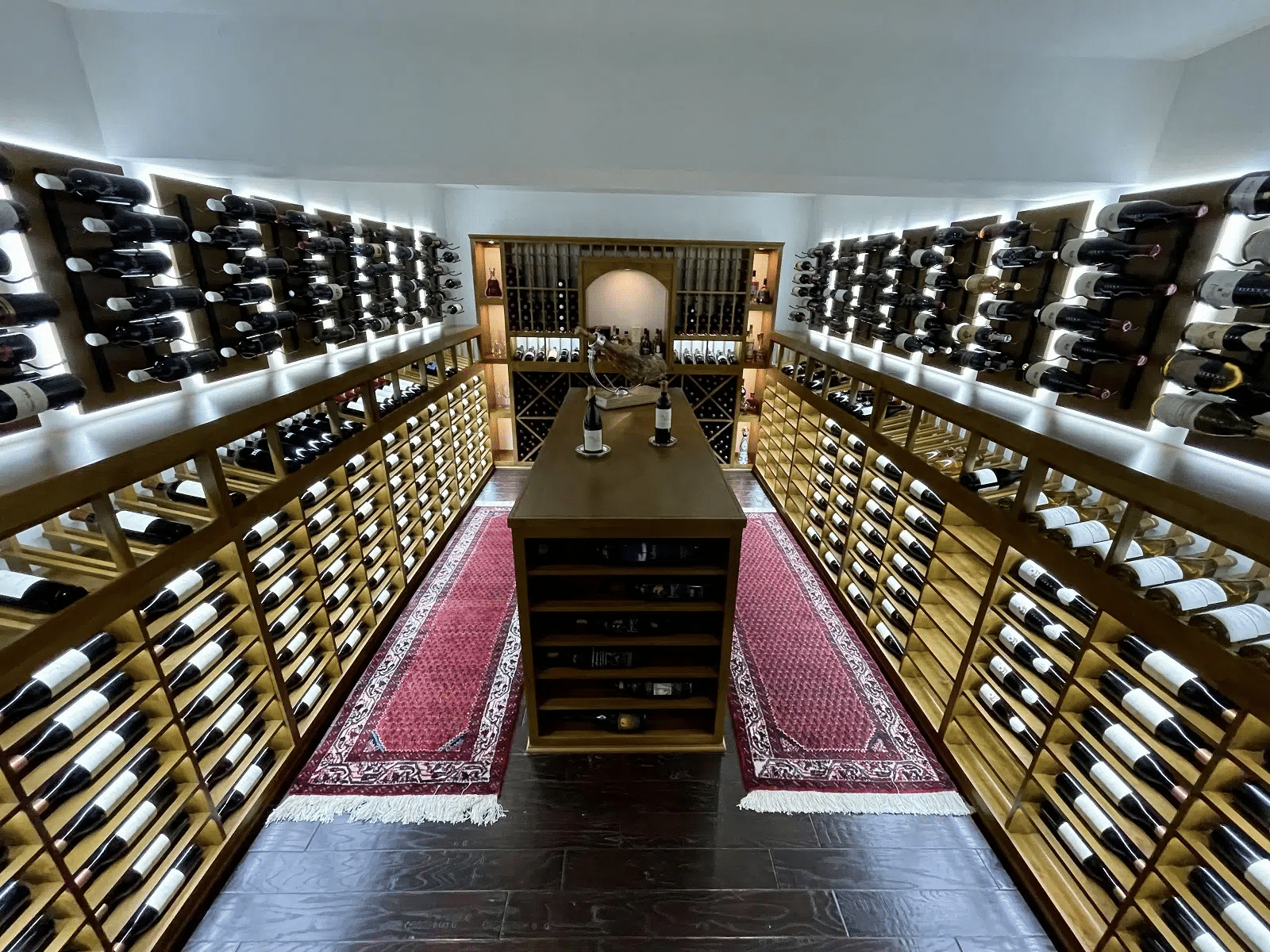 Wine cellar with floor-to-ceiling racks, central table, and red patterned runners under warm lighting