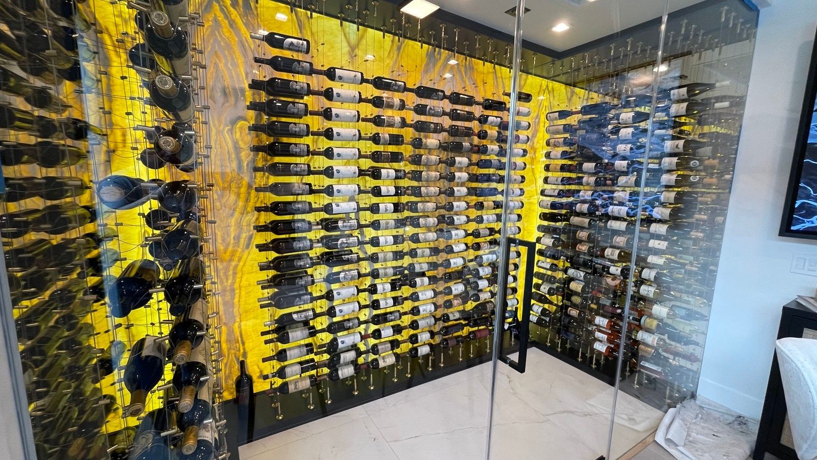 Glass wine cellar with black shelves filled with bottles and yellow accent lighting
