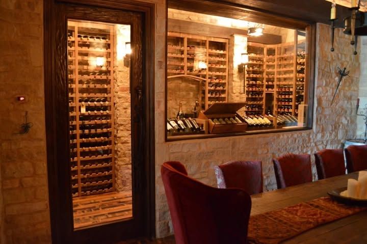 Wine cellar with rows of bottles and a wooden table beside red chairs in warm lighting