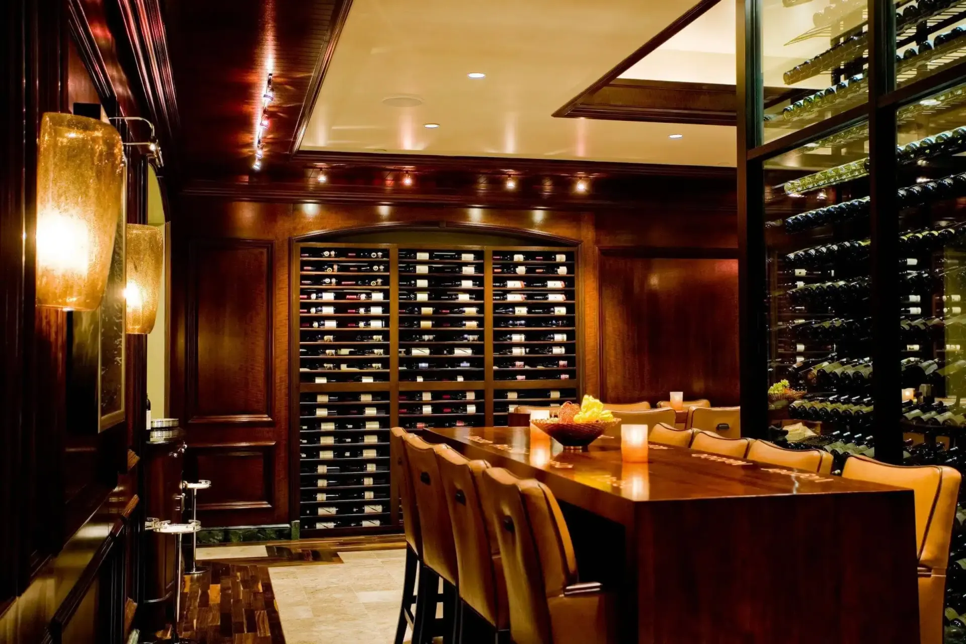 Warmly lit upscale dining room with a long wooden table, leather chairs, and wine storage racks.