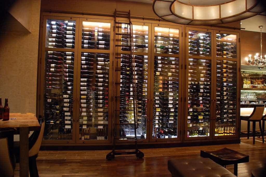 Wine display wall in a warm-lit restaurant interior with shelves of bottles and wooden tables.