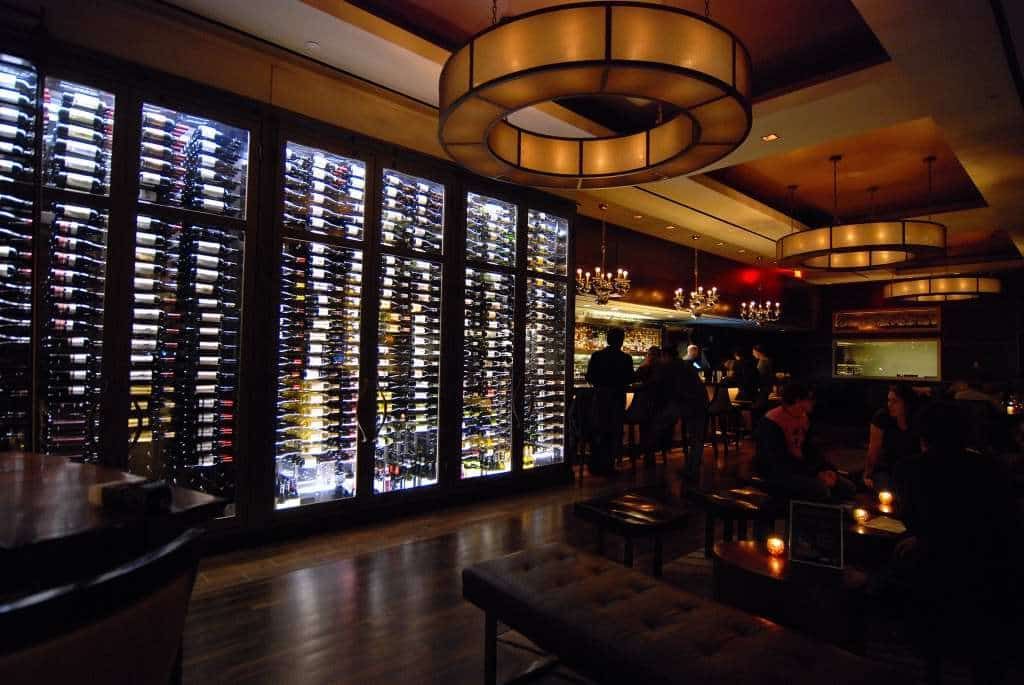 Dimly lit upscale bar with wine wall, hanging lights, and people standing near the counter