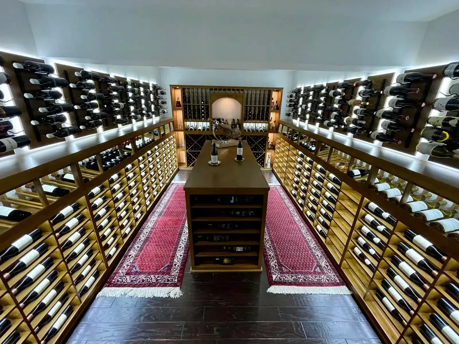 Symmetrical wine cellar with shelves of bottles and a central wooden table over red patterned rugs