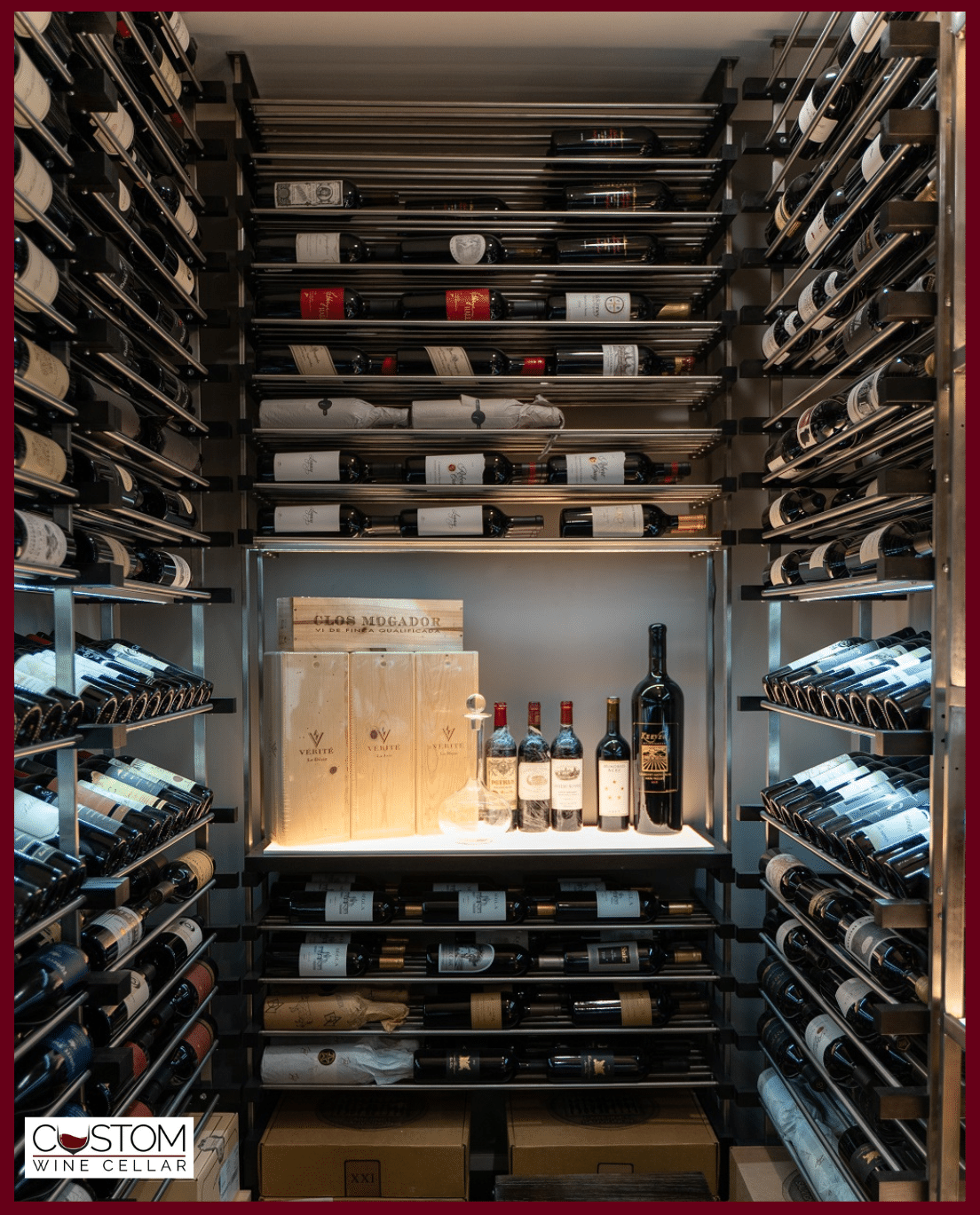 Wall of wine bottles surrounding a lit shelf with boxed bottles and wine glasses in the center