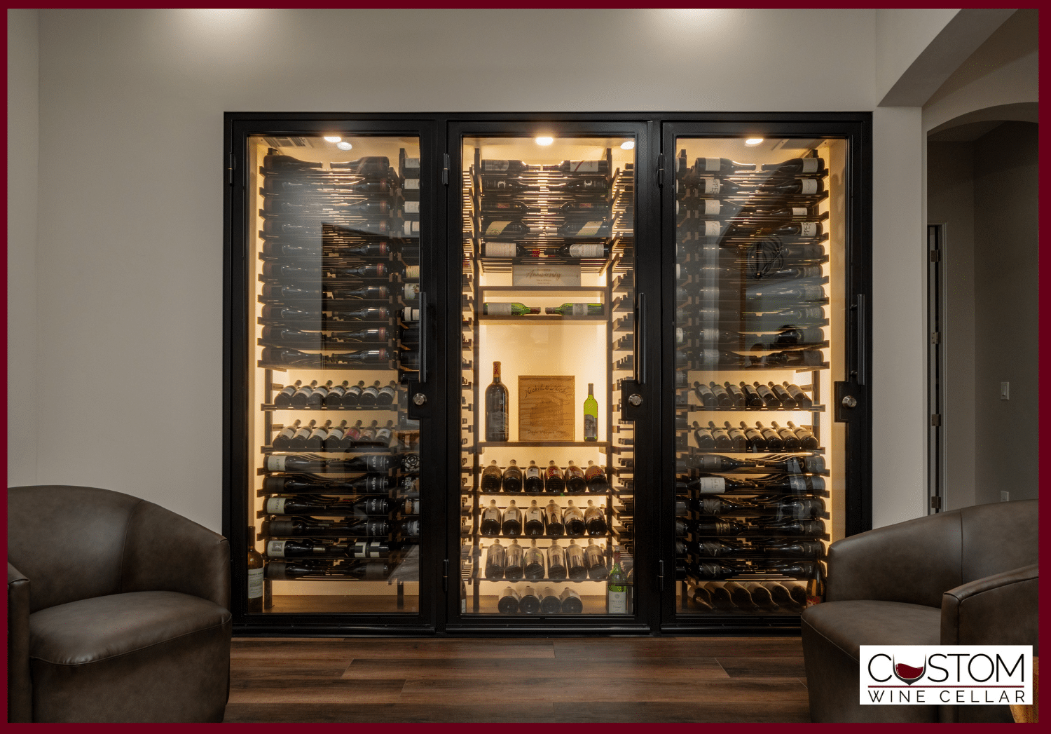 Backlit glass wine cellar with bottles in a modern room, flanked by two armchairs