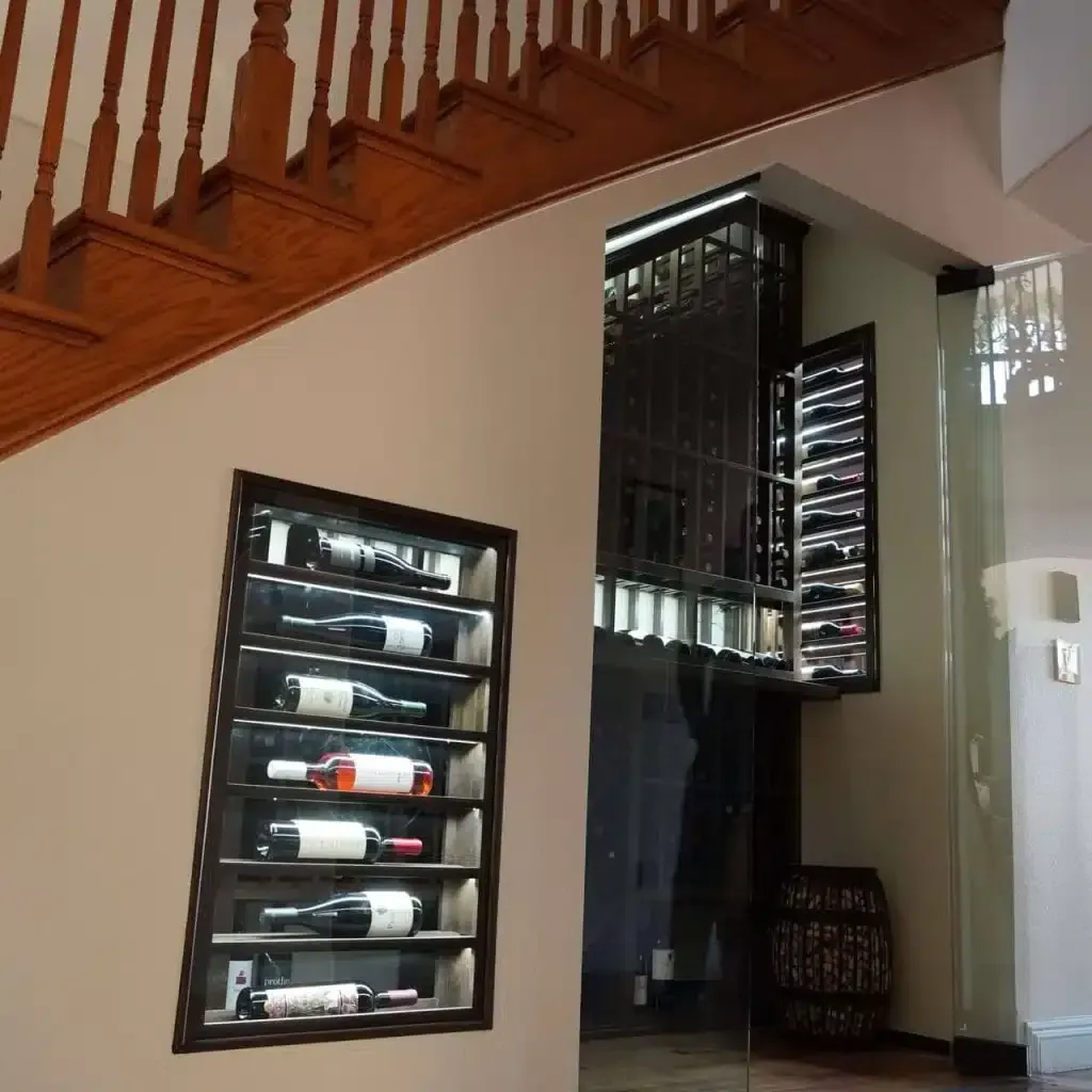 Wine racks under a wooden staircase in a hallway, with bottles displayed on the wall and in a glass cabinet.