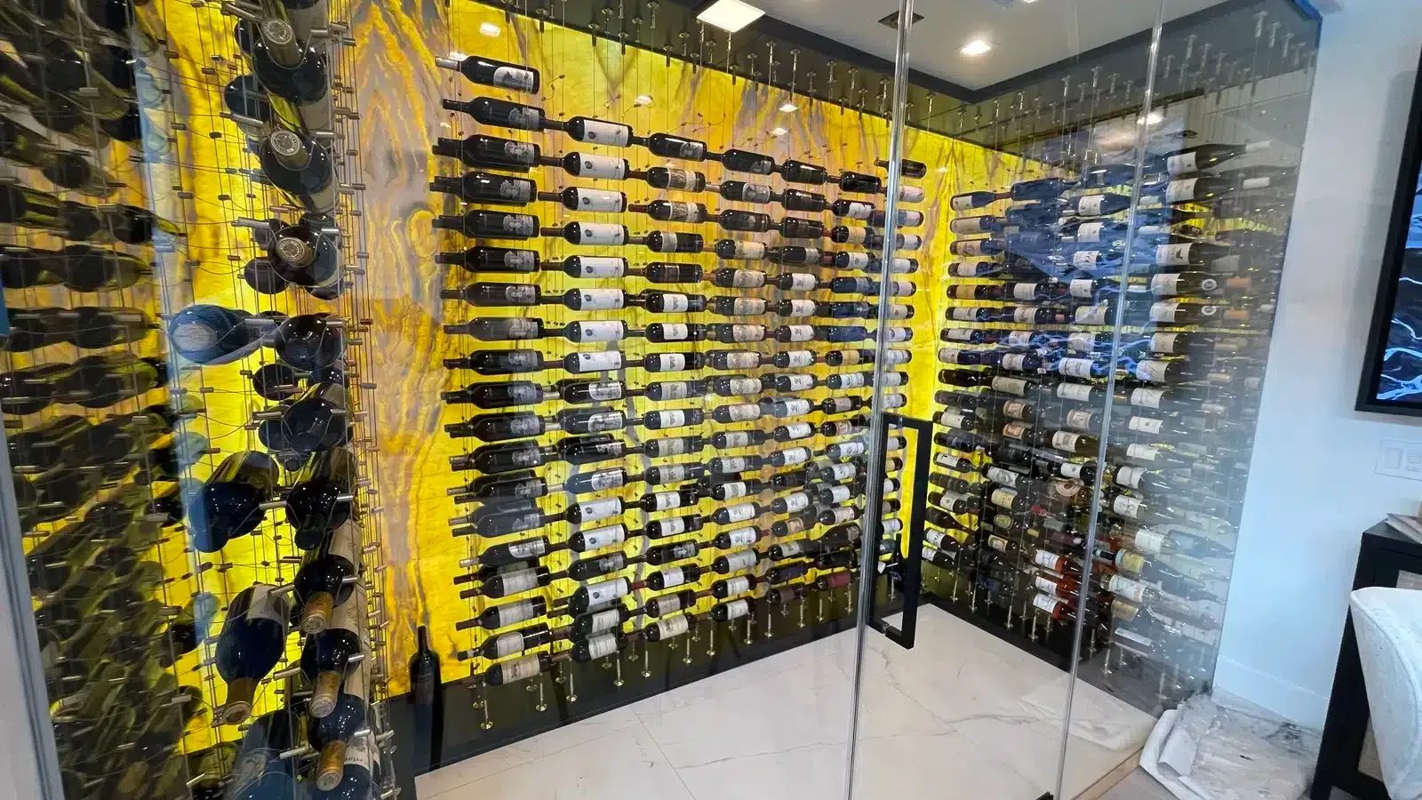 Glass wine cellar with rows of bottles on black racks, highlighted by yellow accent lighting and mirrored walls