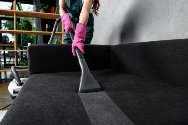 A woman is cleaning a black couch with a vacuum cleaner.