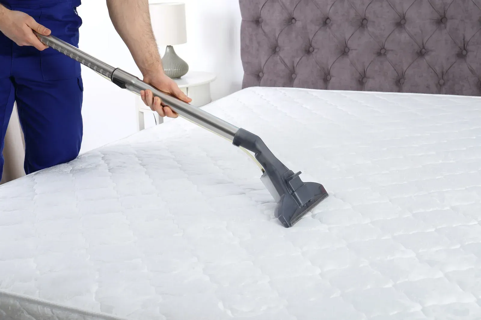 Person vacuuming a white mattress with a cleaning tool in a bedroom.