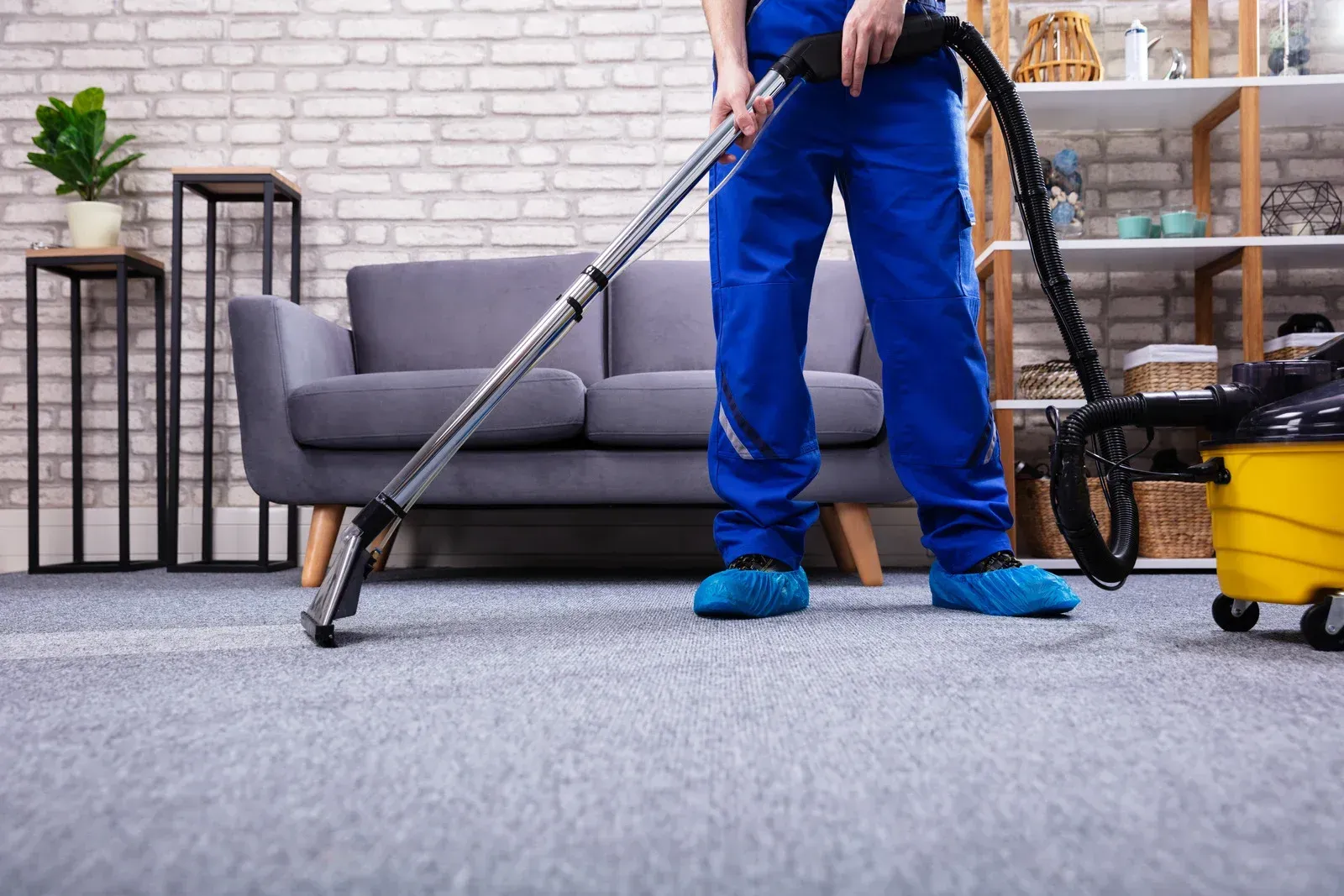 Person in blue cleaning uniform vacuums a gray carpet in a living room.