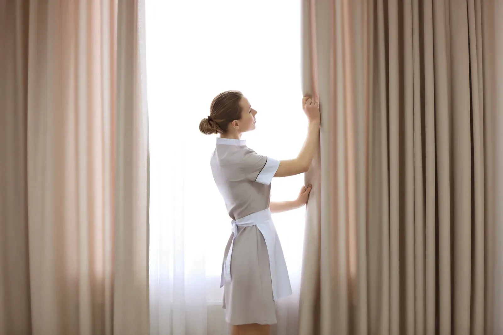 Woman in maid uniform opening curtains in a brightly lit room.