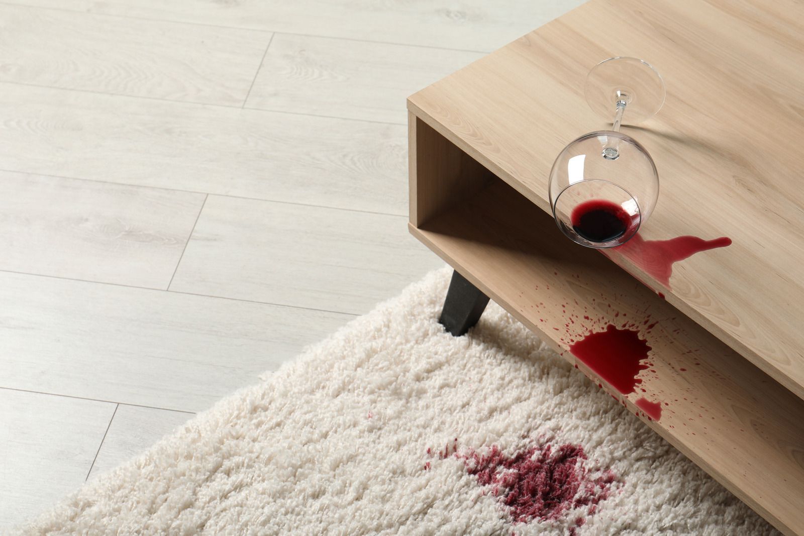 Red wine spills from a toppled glass onto a wooden table and a white rug.