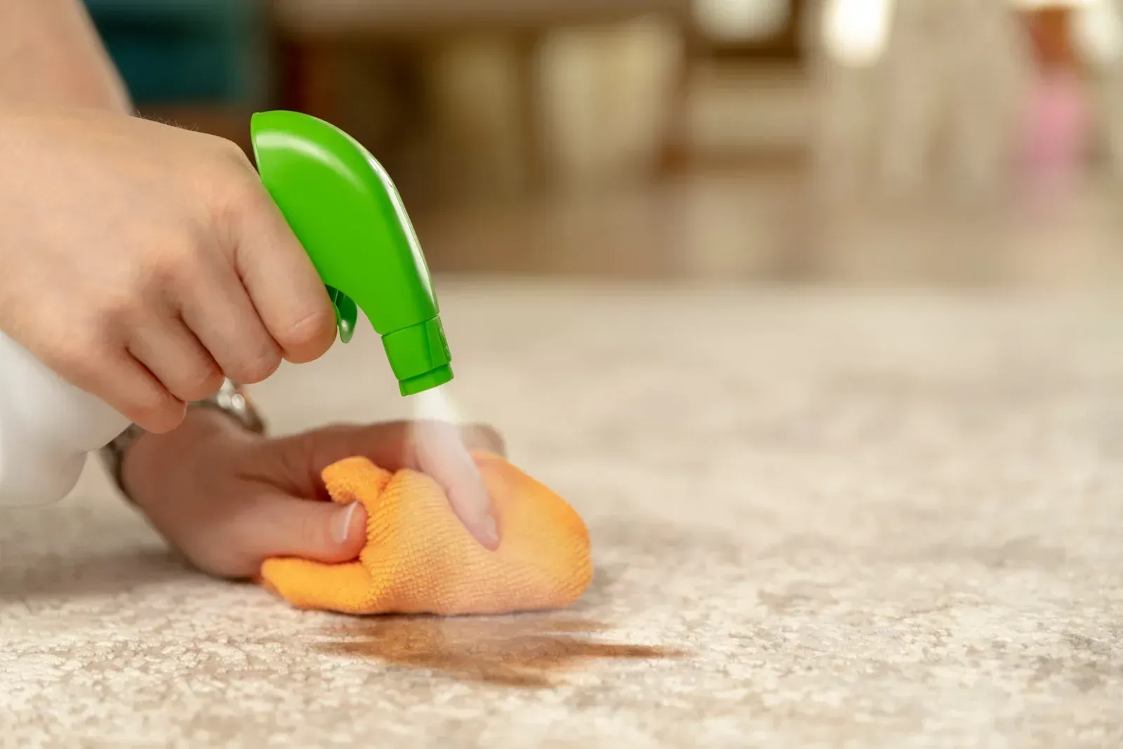 Person spraying a cleaning solution onto a stain on a light-colored surface with an orange cloth.