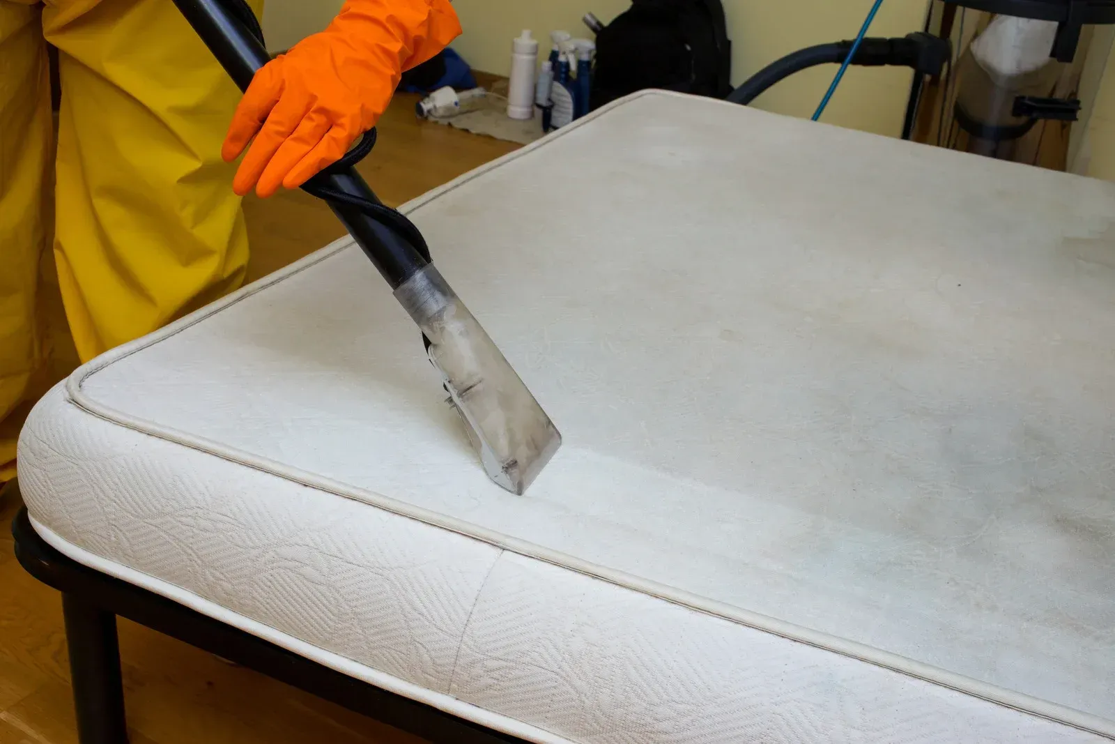 A person wearing orange gloves is cleaning a mattress with a vacuum cleaner.