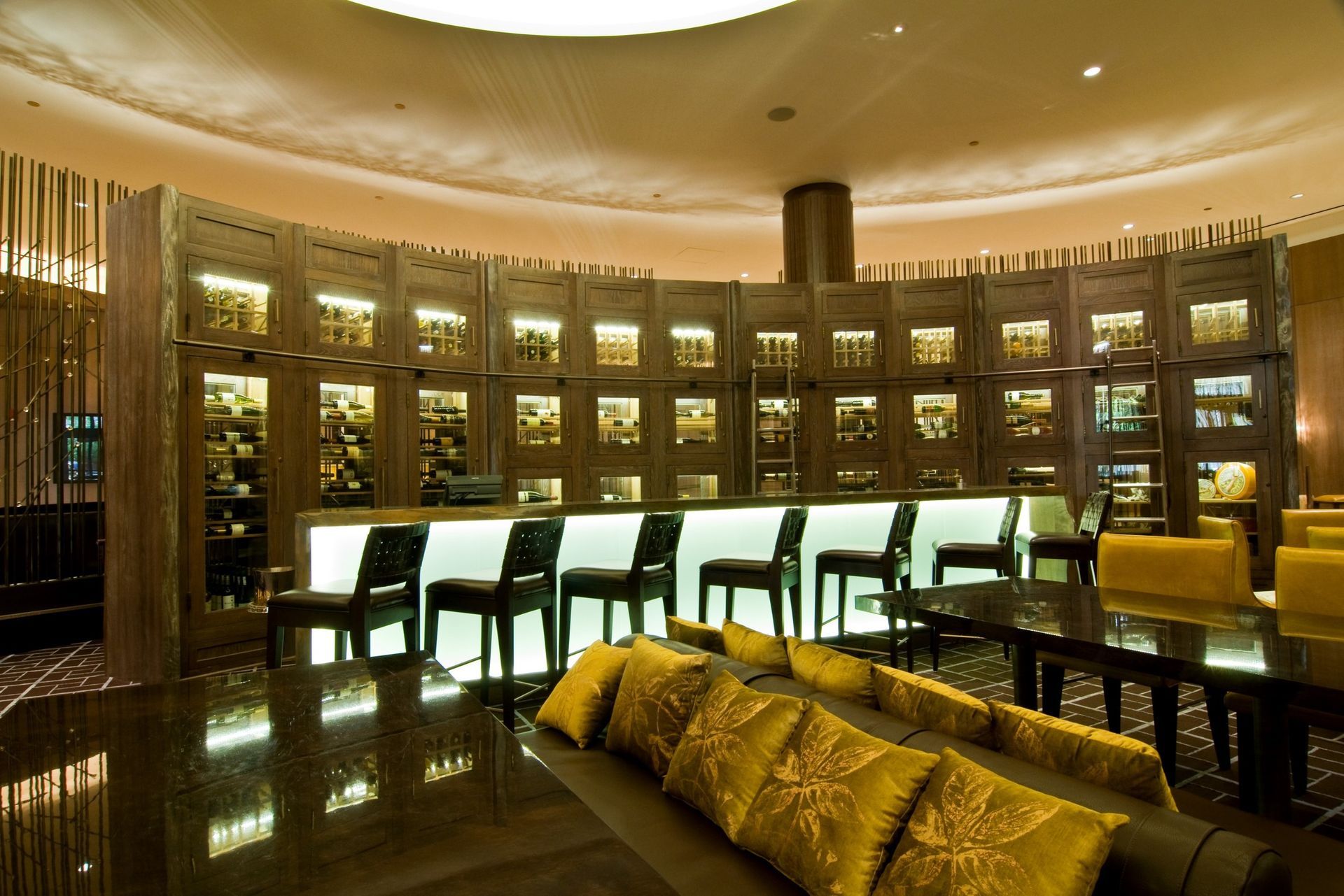 Elegant lounge with curved bar, high stools, dark wood shelves, and gold cushions under warm lighting