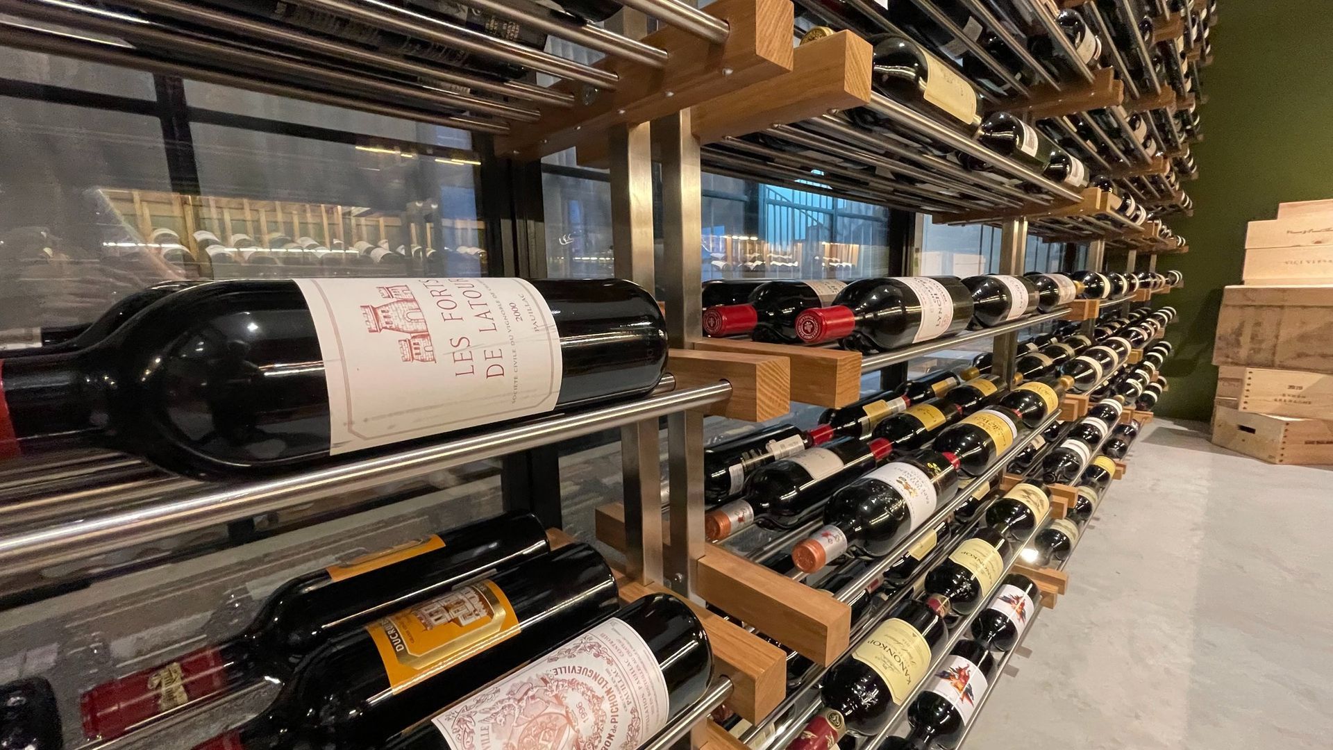 Wine bottles stored on metal racks in a cellar, with rows of labels and a concrete floor.