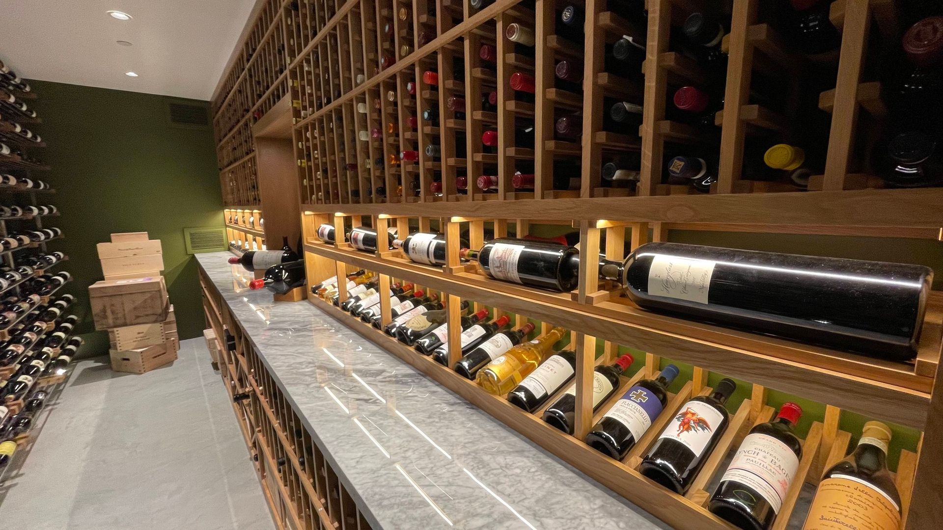 Wine cellar aisle with wooden racks filled with bottles and boxes stacked at the end.