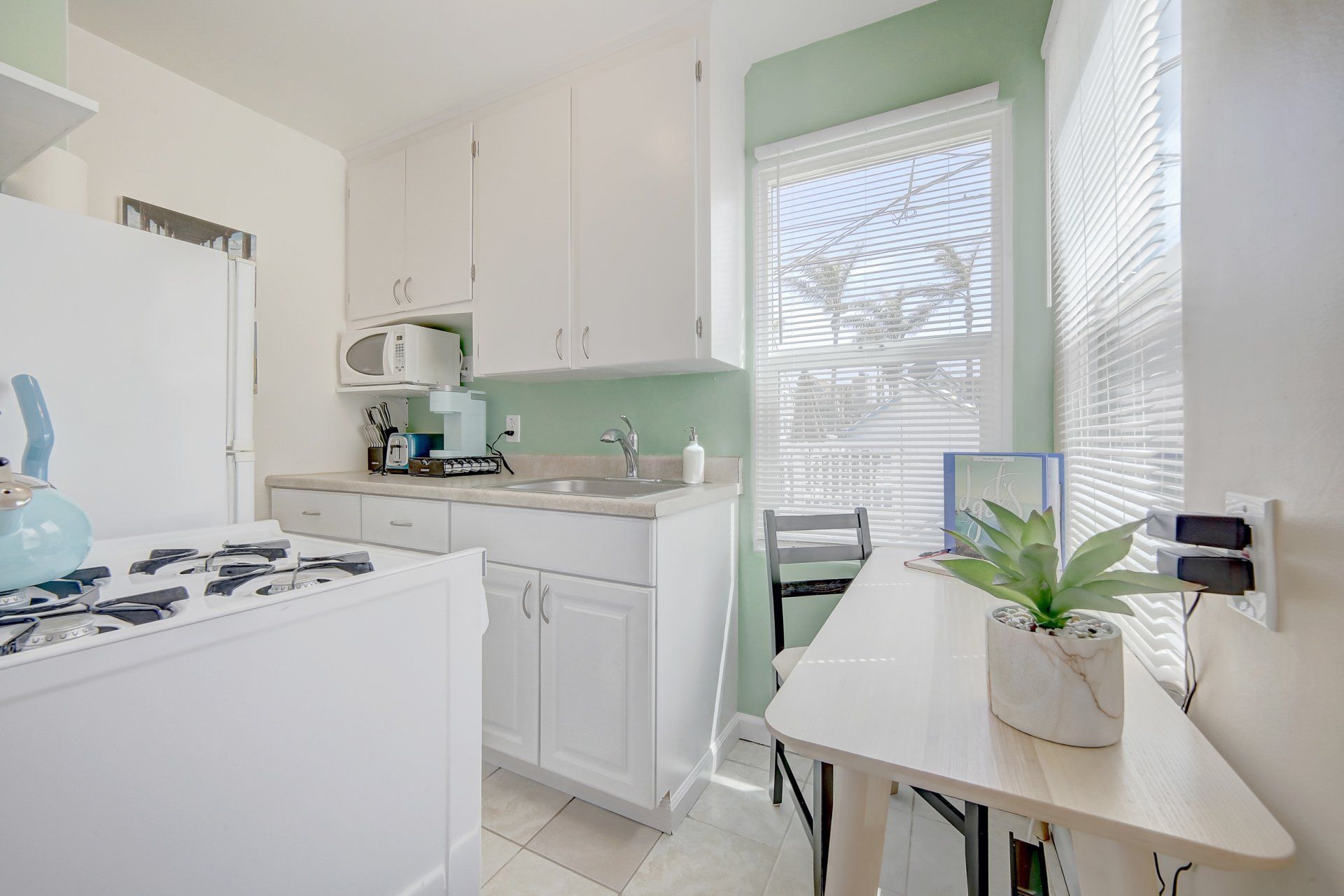 A kitchen with white cabinets , a stove , a desk , a chair and a window.