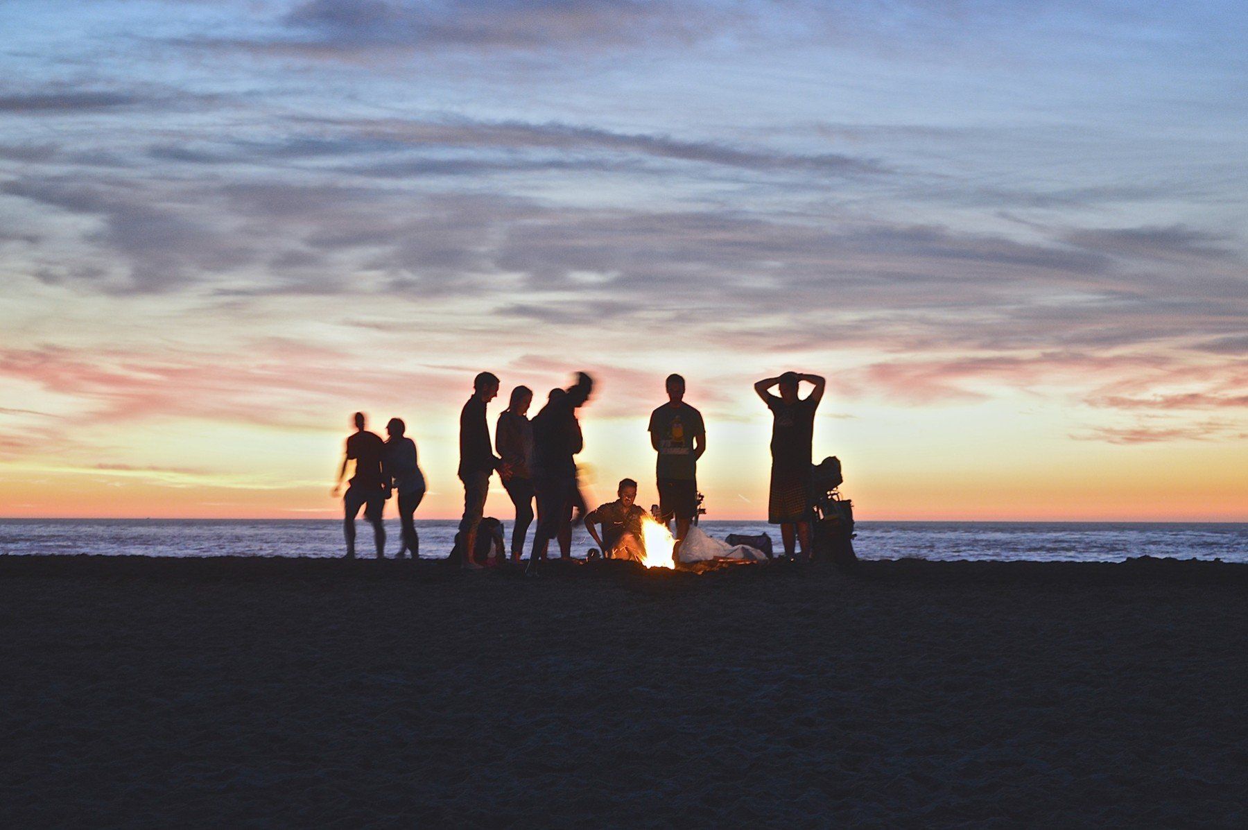 a group of people are standing around a campfire on the beach at sunset .