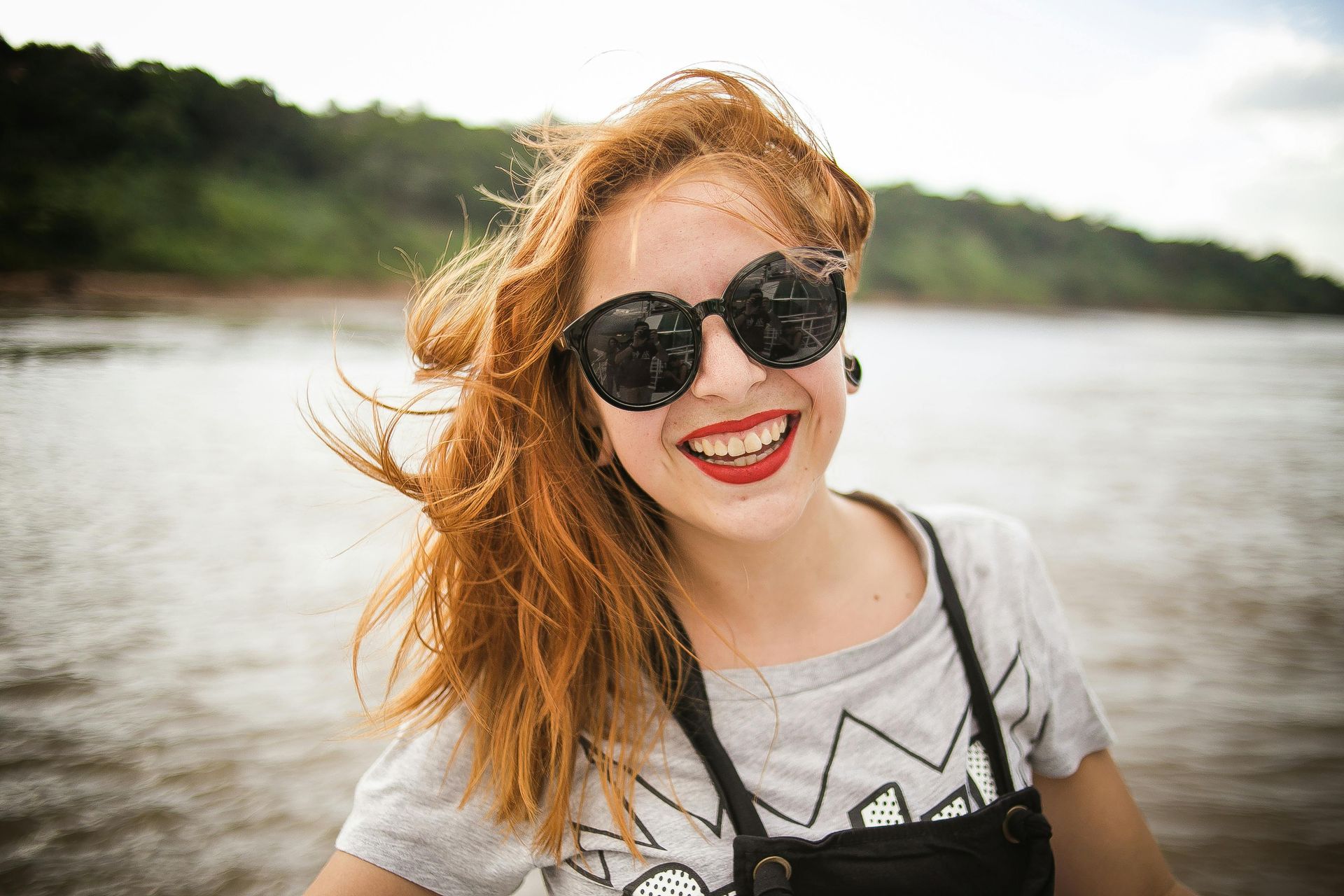 A woman wearing sunglasses and red lipstick is smiling in front of a body of water.