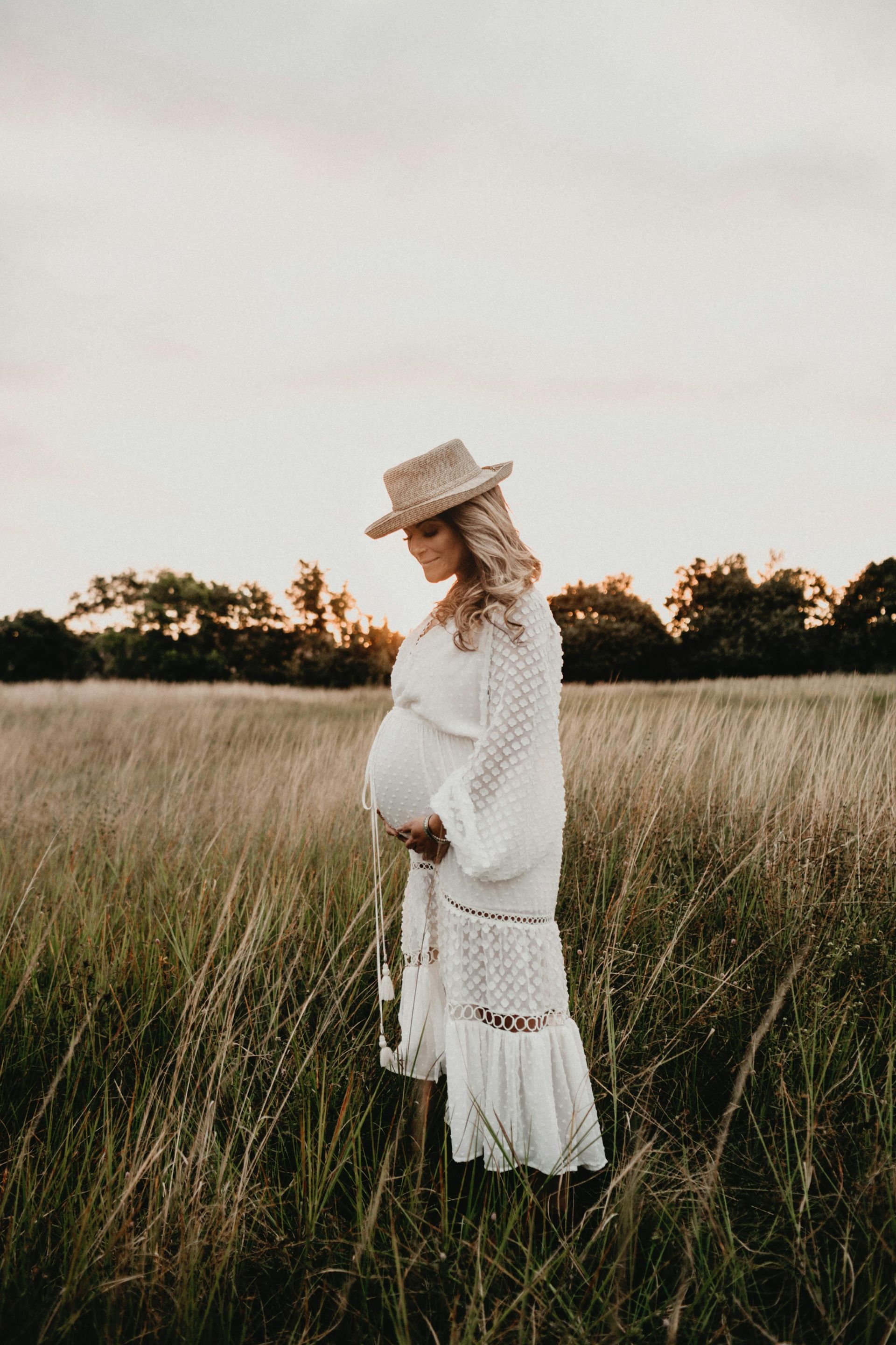 A pregnant woman in a white dress and hat is standing in a field holding her belly.