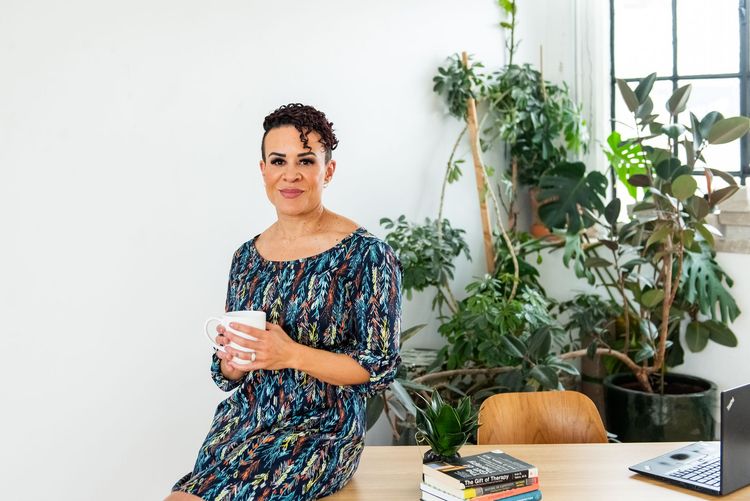 Person in a patterned dress holding a mug beside a laptop and plants in a bright room