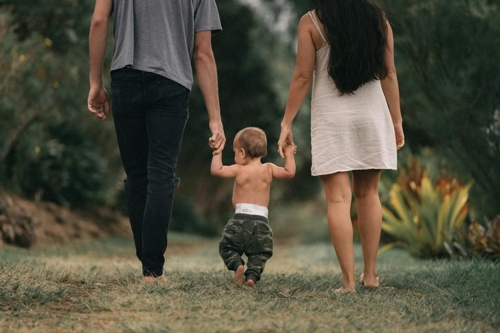 Two adults holding a toddler’s hands while walking barefoot on a grassy path outdoors.