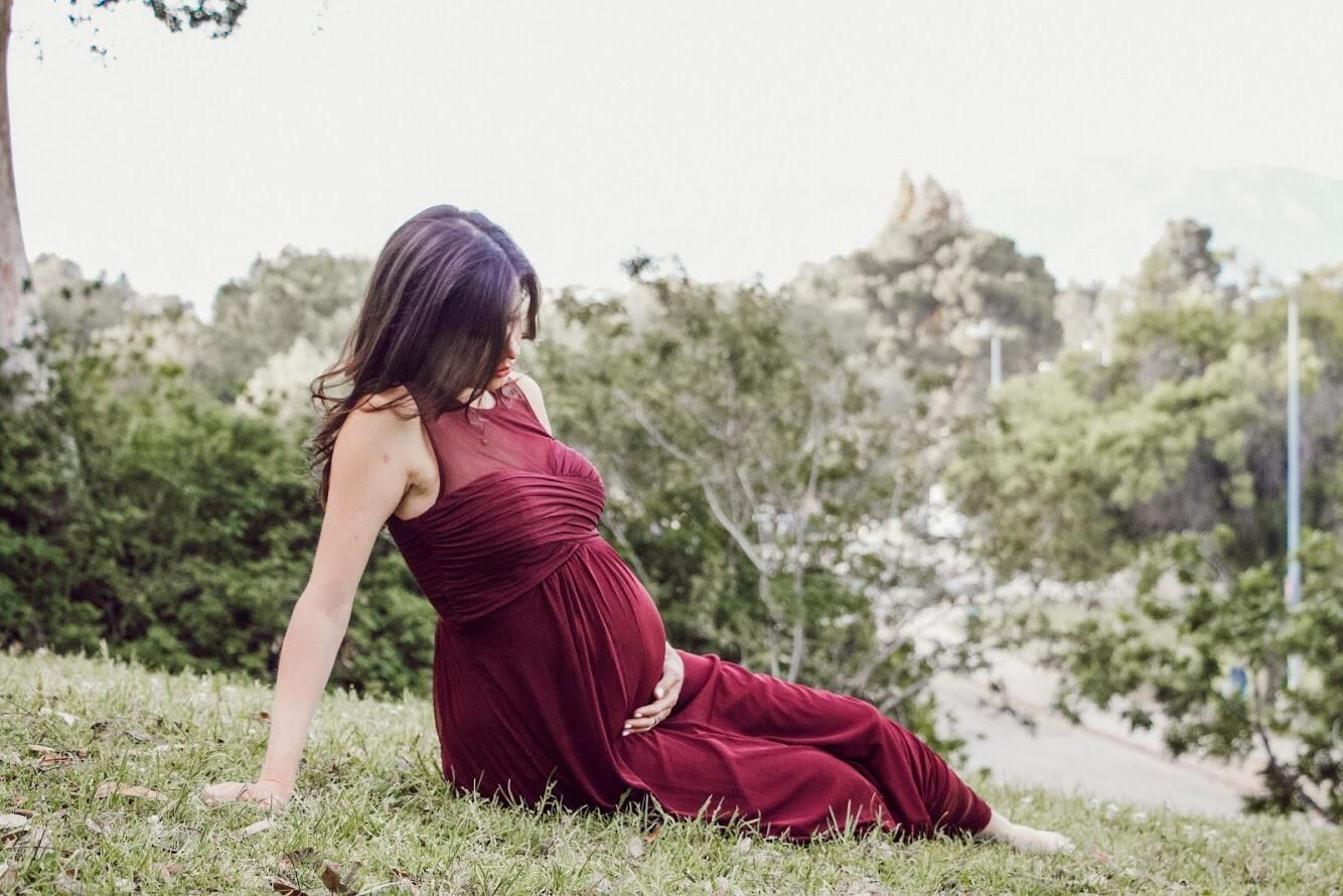 A pregnant woman in a red dress is sitting on the grass holding her belly.
