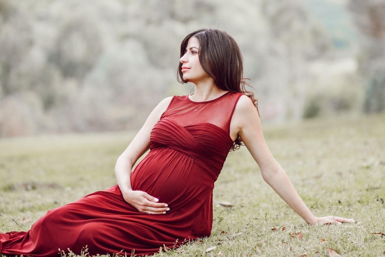 A pregnant woman in a red dress is standing in the grass holding her belly.