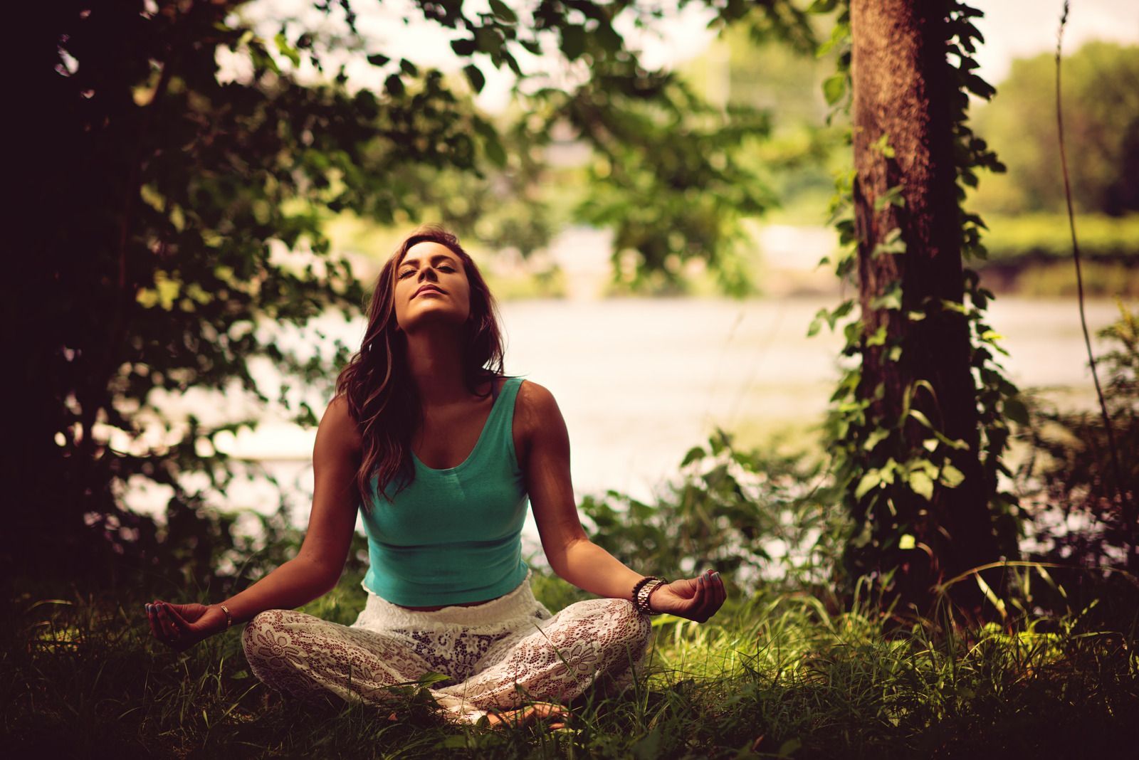 Woman meditating outdoors with arms outstretched, eyes closed. Green top, patterned pants, river in background.