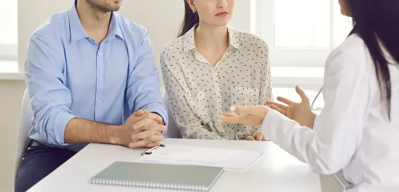 A couple listens intently as a person gestures during a consultation at a table, documents and a notebook visible.