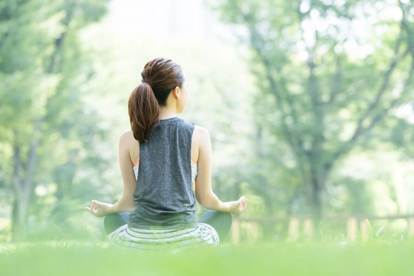 Woman meditating in a park, sitting cross-legged on grass, arms in yoga position, with blurred green background.