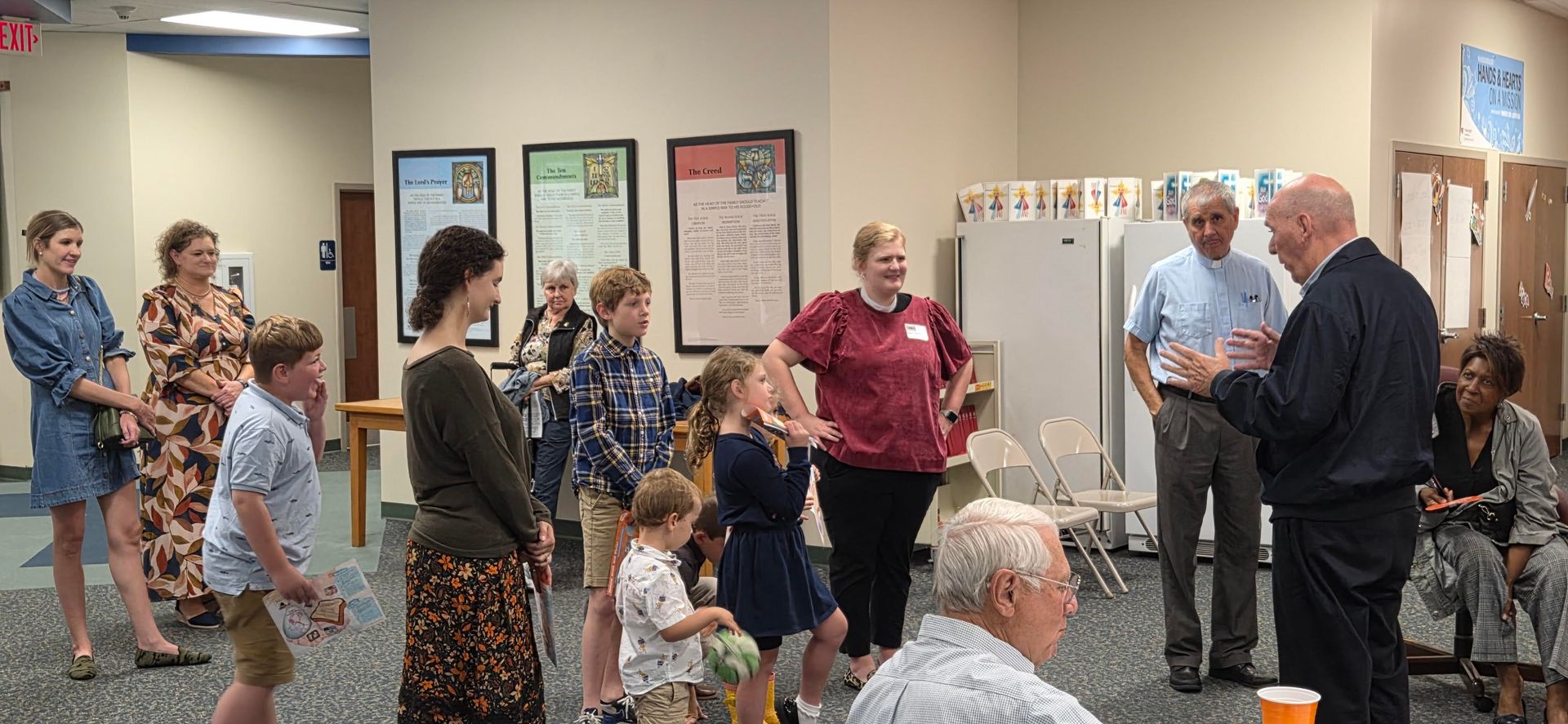 Group of people gathered in a room, listening to a man speaking. Several children and adults standing nearby.