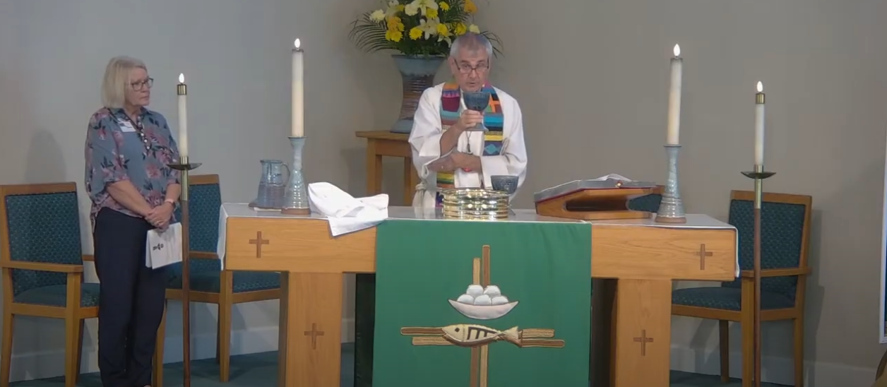 Two people at a church altar with candles, communion items, and flowers. One is speaking.