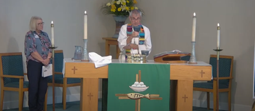 Two people at a church altar with candles, communion items, and flowers. One is speaking.