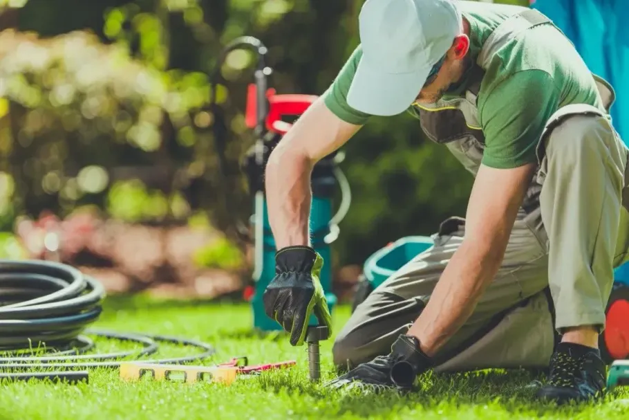 Gardener kneeling on grass, trimming plants with gloves and a hand tool in a sunny yard