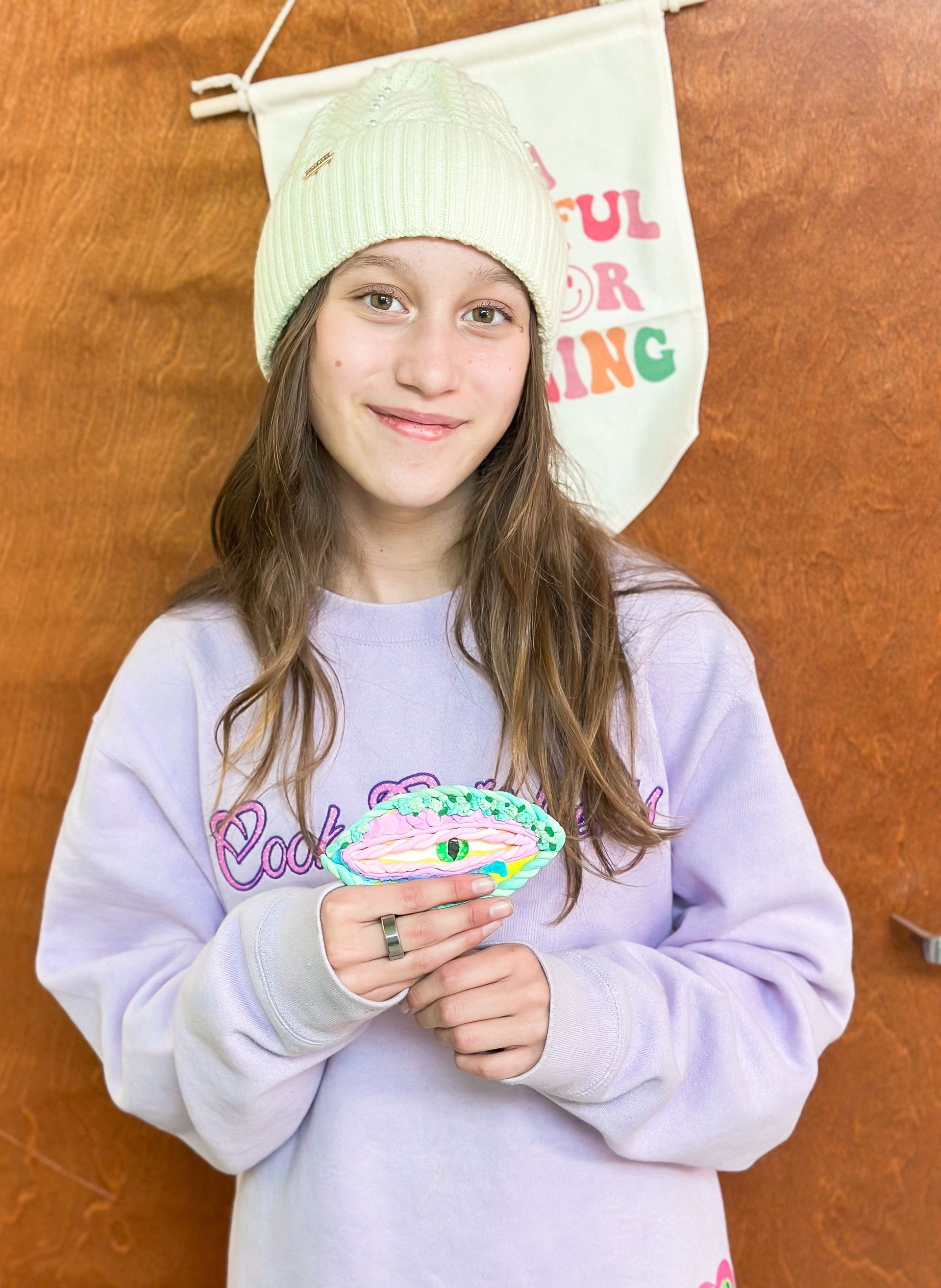 A girl wearing a purple sweatshirt and a white beanie is holding a donut.